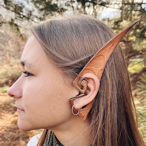 May include: A pair of brown leather elf ears, designed to be worn on the ear. The ears are pointed and have a detailed design.