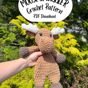 May include: A crocheted moose plushie in shades of brown, with white antlers and blue eyes. The text "MOOSE SNUGGLER Crochet Pattern PDF Download" is displayed above the moose. The plushie is held against a backdrop of green foliage.
