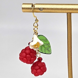 Op de afbeelding: Goudkleurige oorhangers met rode frambozenclusters, witte bloemaccenten en groene blad details. De oorbellen worden getoond op een gouden standaard, die het ontwerp van de sieraden benadrukt. Een charmant accessoire.