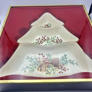 Può includere: Un set di piatti da portata a forma di albero di Natale Lenox. I piatti bianco sporco hanno un bordo dorato e sono decorati con pigne, bacche rosse e foglie verdi. Il set è esposto in una scatola foderata di rosso con una cornice dorata.