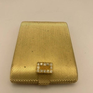 Puede incluir: Un estuche compacto rectangular dorado con un patrón de malla texturizada. El estuche compacto tiene un pequeño cierre rectangular adornado con piedras claras y brillantes. El diseño es vintage y elegante.