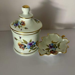 May include: A white porcelain jar with a lid and a matching leaf-shaped dish. Both pieces are decorated with a floral pattern of purple, yellow, and orange pansies with green leaves. The jar and dish have gold trim.