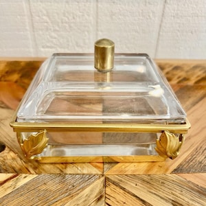 May include: A clear glass and brass serving dish with a square shape and a lid. The dish has a brass handle and decorative leaf accents on the corners. The dish is sitting on a wooden surface.