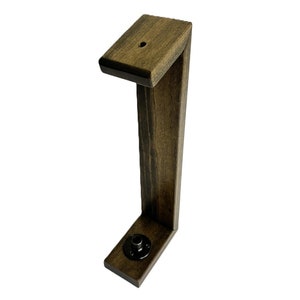 May include: A wooden headphone stand in a dark brown finish. The stand has an L-shape design with a flat base and a top piece with a small hole. A black metal fitting is visible on the base.