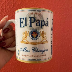 May include: White ceramic mug with a gold rim and handle. The mug features a graphic of a beer label with the text "El Papá" and "Mas Chingon" in blue and black. The label also includes a logo with the text "Cerveceria Modelo Mexico 1925" in gold and red.