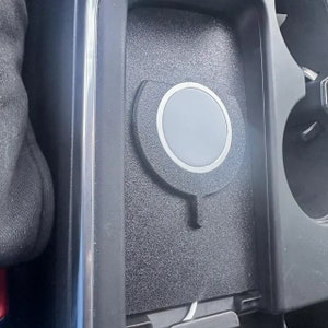 May include: A black textured wireless charging pad with a circular grey centre and a silver metallic ring, integrated into a car centre console storage compartment. The device features a small downward-pointing tab and a white power cable at the base.
