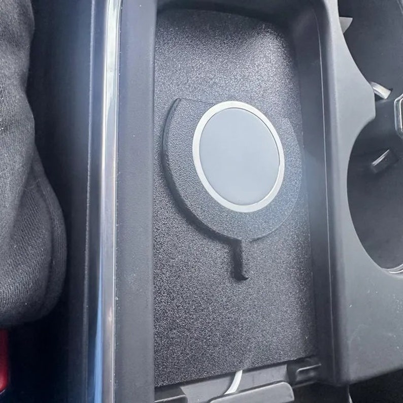 May include: A black textured wireless charging pad with a circular grey centre and a silver metallic ring, integrated into a car centre console storage compartment. The device features a small downward-pointing tab and a white power cable at the base.