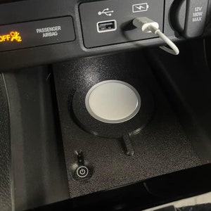 May include: A black textured wireless charging pad installed in a vehicle centre console. The circular white charging disc is centred on the base, which features a power button with a circular icon. A white USB cable is plugged into the port above.