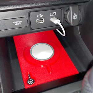 May include: A red wireless charging pad insert designed for a vehicle centre console, featuring a circular white charging surface, a power button icon, and a white charging cable plugged into the USB port above the console storage area.