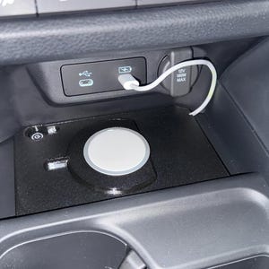 May include: A black wireless charging pad installed in a vehicle centre console storage area. The circular white charger is connected by a white cable to a USB port located on the dashboard panel above, which also features a 12V power outlet.