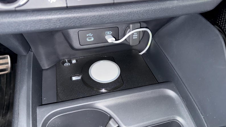 May include: A black wireless charging pad installed in a vehicle centre console storage area. The circular white charger is connected by a white cable to a USB port located on the dashboard panel above, which also features a 12V power outlet.