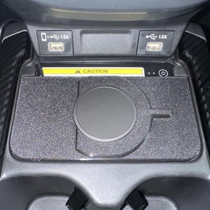 May include: A black wireless charging pad installed in a vehicle centre console. The rectangular pad features a circular charging area and a power button icon. Above the pad are two USB ports labelled 1.5A and 1.0A, and a yellow caution warning label.