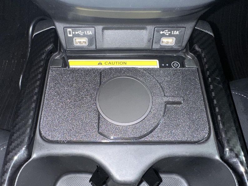 May include: A black wireless charging pad installed in a vehicle centre console. The rectangular pad features a circular charging area and a power button icon. Above the pad are two USB ports labelled 1.5A and 1.0A, and a yellow caution warning label.