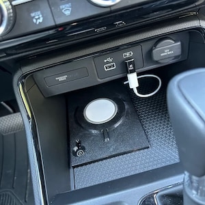 May include: A black wireless charging pad with a circular white centre and a power button icon, installed in a car centre console. A white USB-C cable is plugged into a black adapter in the vehicle's USB port, with the text 40 Gbps visible.