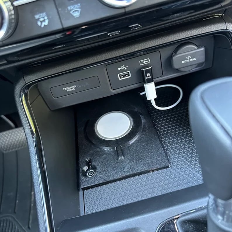 May include: A black wireless charging pad with a circular white centre and a power button icon, installed in a car centre console. A white USB-C cable is plugged into a black adapter in the vehicle's USB port, with the text 40 Gbps visible.