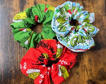 Puede incluir: Tres coleteros de tela con diseños temáticos de Grinch. Uno es verde con diseños de Grinch y copos de nieve, otro es rojo con diseños de Grinch y copos de nieve, y el tercero es blanco y azul con diseños de Grinch.
