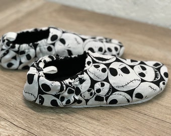 Może przedstawiać: Para czarno-białych bucików dla niemowląt. Buty mają powtarzający się wzór twarzy Jacka Skellingtona z Miasteczka Halloween. Wnętrze jest czarne, a podeszwy białe.