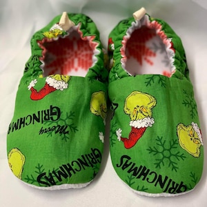 以下が含まれることがあります： 緑色のGrinchをテーマにしたベビー用スリッパのペア。スリッパには、Grinchの顔、雪の結晶、そして「Merry Grinchmas」という言葉の繰り返し模様があります。内側は白で、赤と白の模様です。