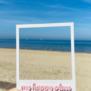Könnte beinhalten: Ein weißer, quadratischer Fotorahmen mit einem Rand, der unten den Schriftzug "my happy place" in einer rosa Schreibschrift trägt. Der Rahmen wird vor einen Sandstrand, das Meer und einen klaren blauen Himmel gehalten.