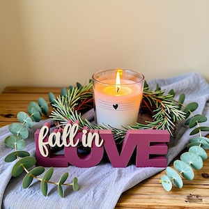 Könnte beinhalten: Eine brennende Kerze in einem Glas mit einem kleinen schwarzen Herz und horizontalen Linien, platziert in einem grünen Tannenkranz. Vorne zeigt ein dunkelrosa Holzschild den Text "fall in LOVE" mit einem gewundenen Eukalyptuszweig.