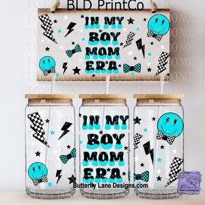 May include: Three clear glass tumblers with bamboo lids, each featuring a design with the text "IN MY BOY MOM ERA" in turquoise and black. The design includes lightning bolts, stars, and smiley faces. A wooden sign above repeats the design.