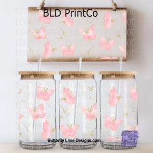 May include: Three clear glass tumblers with bamboo lids and straws, decorated with pink butterflies and gold accents. A wooden sign above displays the text "BLD PrintCo" and a butterfly pattern. The text "Butterfly Lane Designs.com" is also visible.