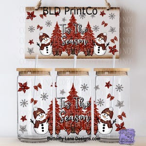 May include: Three glass tumblers with bamboo lids and straws, featuring a festive winter design. The design includes snowmen, Christmas trees, bows, snowflakes, and the text "Tis The Season". A wooden sign with the same design hangs above the tumblers.