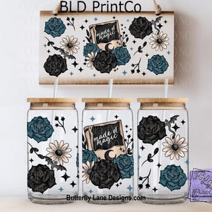 May include: A set of three glass tumblers with wooden lids, featuring a floral and bat design. The design includes black and teal roses, daisies, and the text "made of magic". A matching sign hangs above the tumblers.