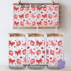 May include: Three glass tumblers with bamboo lids and straws, decorated with a pattern of red strawberries, bows, and hearts on a white background. A matching sign hangs above the tumblers, also displaying the strawberry design.