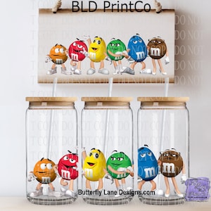 May include: Clear glass tumblers with bamboo lids and straws, each featuring a different color M&M character. The characters are orange, red, yellow, green, blue, and brown. A sign above reads "BLD PrintCo". The website "Butterfly Lane Designs.com" is also visible.