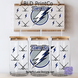 May include: Clear glass tumblers with wooden lids and straws, featuring the Tampa Bay Lightning logo, hockey sticks, and lightning bolts. A wooden sign above displays the team logo and text. The text "Butterfly Lane Designs.com" is visible.