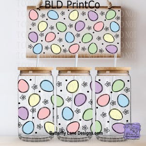 May include: Three glass tumblers with wooden lids and a matching sign, all decorated with a pastel Easter egg pattern. The sign and tumblers feature eggs in pink, yellow, blue, green, and purple, with small black flower accents. The text "Butterfly Lane Designs.com" is visible.
