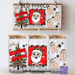 May include: Clear glass tumblers with bamboo lids feature Christmas designs. One has a leopard print Christmas tree with "Rocking Around" text. Another has Santa Claus with a leopard print hat. The third has gingerbread men and stars.