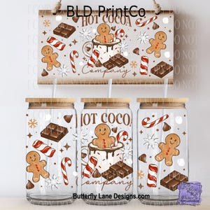 May include: Three clear glass tumblers with bamboo lids and straws, decorated with a "Hot Cocoa Company" design. The design includes gingerbread men, candy canes, chocolate bars, and snowflakes. A matching sign hangs above.