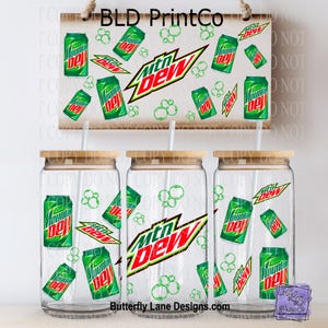 May include: Clear glass tumblers with bamboo lids and straws, featuring a Mountain Dew design. The design includes green soda cans, the Mountain Dew logo, and bubbles. A wooden sign with the text "BLD PrintCo" is visible.