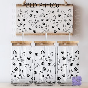 May include: Three clear glass tumblers with bamboo lids and straws, decorated with a repeating pattern of black cat faces and paw prints. A wooden sign above displays the same design. The sign reads "BLD PrintCo".