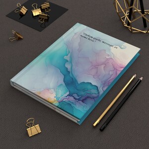 May include: A teal blue journal with a watercolor design of blue, purple, and pink abstract shapes. The journal has a white spine and a white interior. The text "Custom quote, message, or name here." is printed on the front cover.
