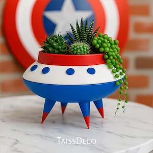 Könnte beinhalten: Ein neuartiger Pflanztopf in Form eines Retro-Raumschiffs, bemalt in Blau, Weiß und Rot, mit blauen Bullaugen-Details und rotspitzigen Beinen. Der Pflanztopf enthält eine Vielzahl von grünen Sukkulenten und Kakteen. Der Text "TaissDeco" ist sichtbar.