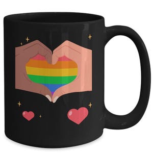 May include: Black ceramic mug featuring a heart formed by two hands with diverse skin tones, enclosing a rainbow-colored heart. Two pink hearts and starbursts accent the design. A perfect gift for anyone.
