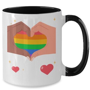 May include: White ceramic mug with a black interior and handle. The mug features a heart made of rainbow stripes, formed by two hands. Two red hearts and gold starbursts are also on the mug.