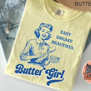 Pode incluir: Uma camiseta amarela com uma ilustra&ccedil;&atilde;o retr&ocirc; de uma mulher segurando uma faca de manteiga e uma barra de manteiga. O texto "Butter Girl" est&aacute; escrito em uma fonte retr&ocirc; abaixo da ilustra&ccedil;&atilde;o. O texto "Easy Greasy Beautiful" est&aacute; escrito em azul acima da ilustra&ccedil;&atilde;o.