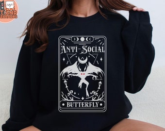 Puede incluir: Sudadera negra con un gráfico blanco de una carta de tarot. La carta tiene una mariposa blanca con ojos rojos y el texto "Anti-Social Butterfly".