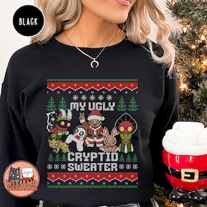 May include: Black crew neck sweatshirt with a festive design. The design features the text "MY UGLY CRYPTID SWEATER" with cartoon cryptids and Christmas trees in red, green, and white. The brand logo "Wit and Wisecracks" is in the bottom left corner.