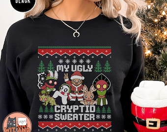 May include: Black crew neck sweatshirt with a festive design. The design features the text "MY UGLY CRYPTID SWEATER" with cartoon cryptids and Christmas trees in red, green, and white. The brand logo "Wit and Wisecracks" is in the bottom left corner.