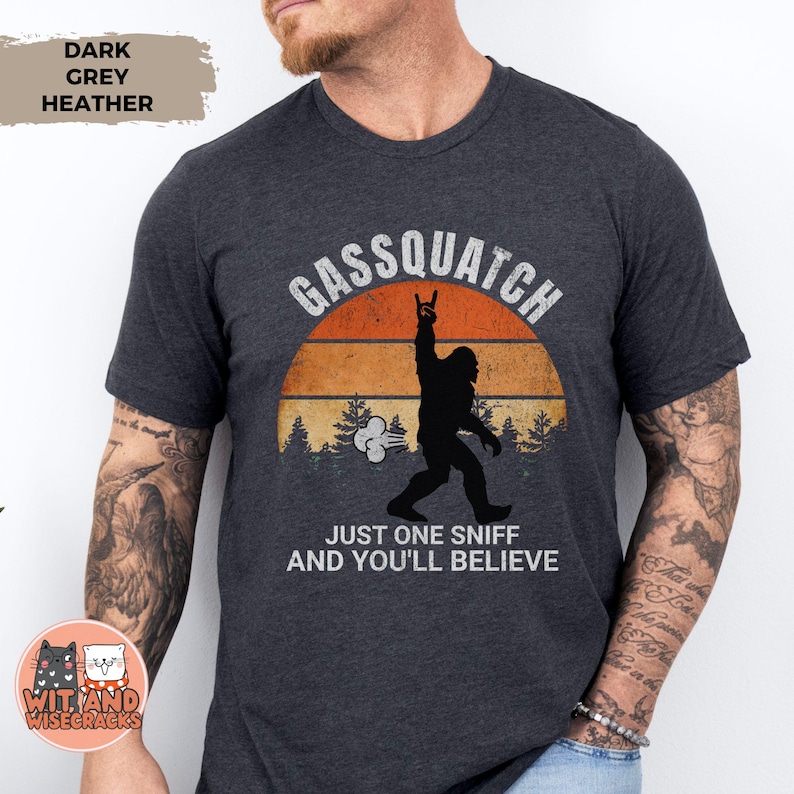 May include: Dark grey heather t-shirt with the text "Gassquatch" above a silhouette of Bigfoot giving a rock on hand gesture. The text "Just one sniff and you'll believe" is below the graphic. The shirt has a retro sunset design.