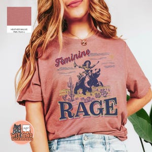 May include: A heather mauve t-shirt with a graphic of a woman riding a bear and the text "Feminine Rage".