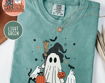 May include: Light green Comfort Colors t-shirt featuring a Halloween ghost design. The graphic includes a ghost holding a broom and pumpkin, with bats and stars. The shirt has a crew neck and short sleeves. The brand name "Wit and Wisecracks" is visible.