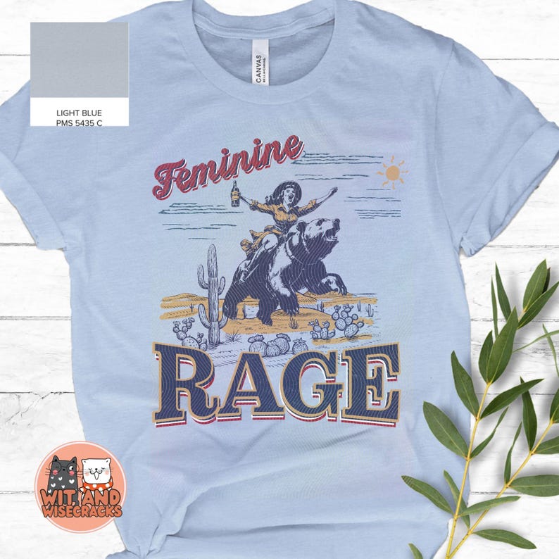 May include: Light blue t-shirt with a vintage-style graphic of a woman riding a bear. The graphic includes the text "Feminine Rage".