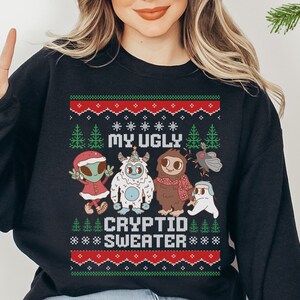 Könnte beinhalten: Schwarzes Sweatshirt mit Rundhalsausschnitt und festlichem Design. Die Grafik zeigt den Text "MY UGLY CRYPTID SWEATER" über Cartoon-Kryptiden wie einem Alien, Yeti, Bigfoot, Mothman und einem Geist. Das Design beinhaltet Weihnachtsbäume und Schneeflocken.