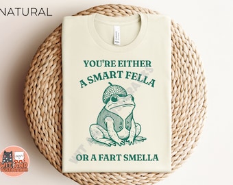 May include: A white t-shirt with a green frog wearing a hat and the text "You're either a smart fella or a fart smella".
